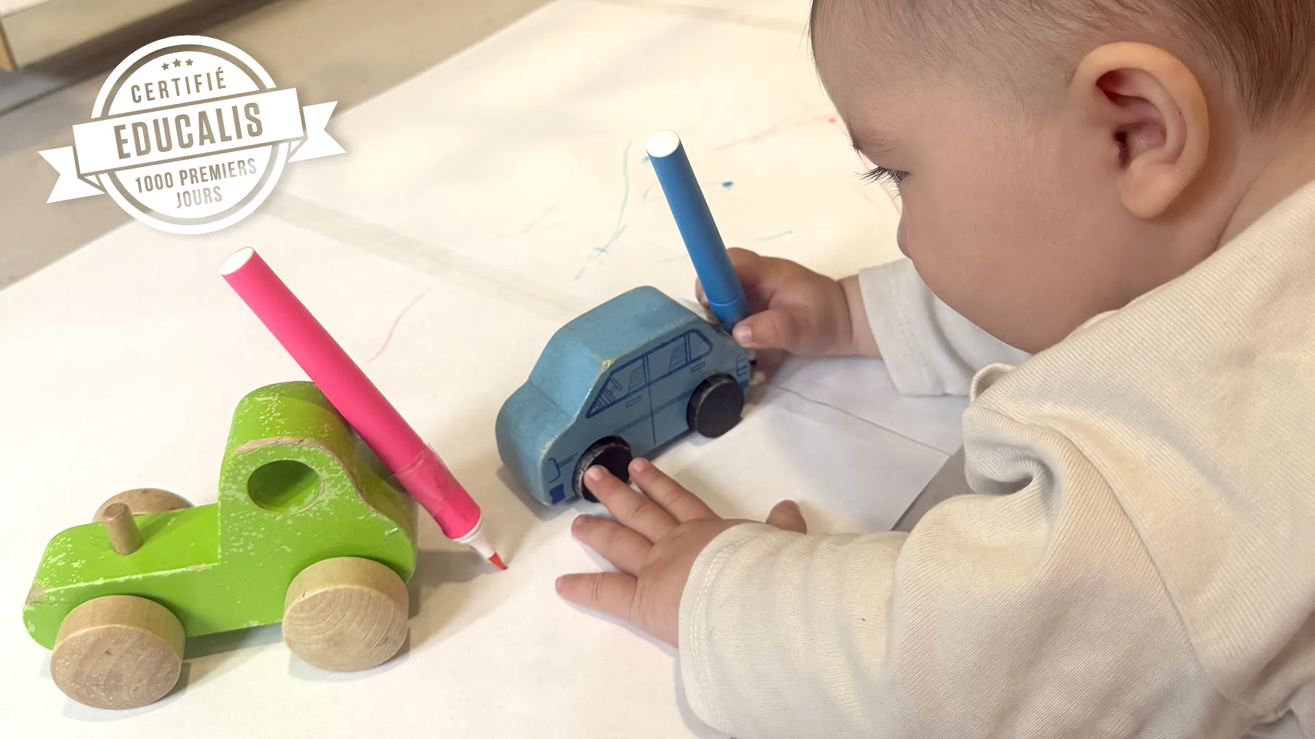 Un enfant à la crèche La Nanosphère à Lausanne dessine avec une voiture équipée d’un feutre sur une grande feuille au sol.